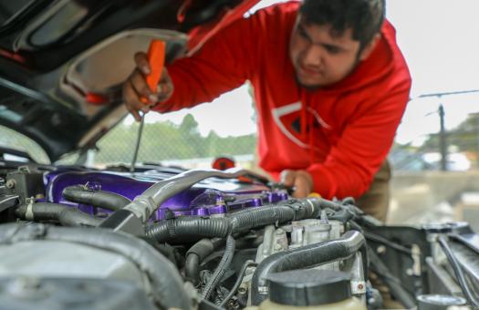 student in red hoodie leaning over car and looking at engine with screwdriver in hand