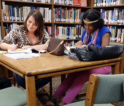two female students studying at a library table with stacks of books around them