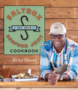 Chef Ricky Moore standing next to a sign for the Saltbox Seafood Joint Cookbook