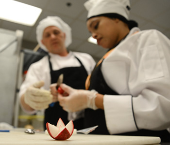 culinary arts student in chef whites and apron practices carving a radish into a flower shape