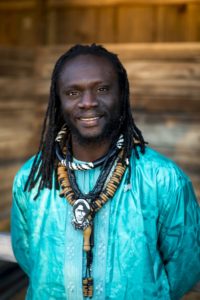 Diali Cissokho wearing a colorful blue shirt and heavy bead necklaces, sporting dreadlocks