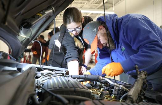 Two students looking under the hood of a car with a flashlight to diagnose a problem